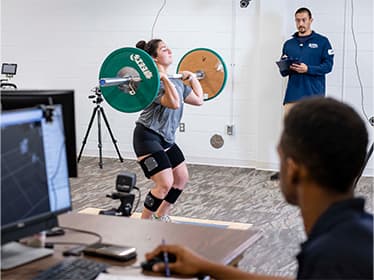 For Holistic Fitness & Wellness research, two Georgia Southern students assess the strength of an athlete holding a barbell.