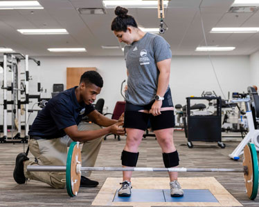 A student from Georgia Southern's Waters College of Health Professions preps a test subject standing in front of a set of barbells in an athletic training setting