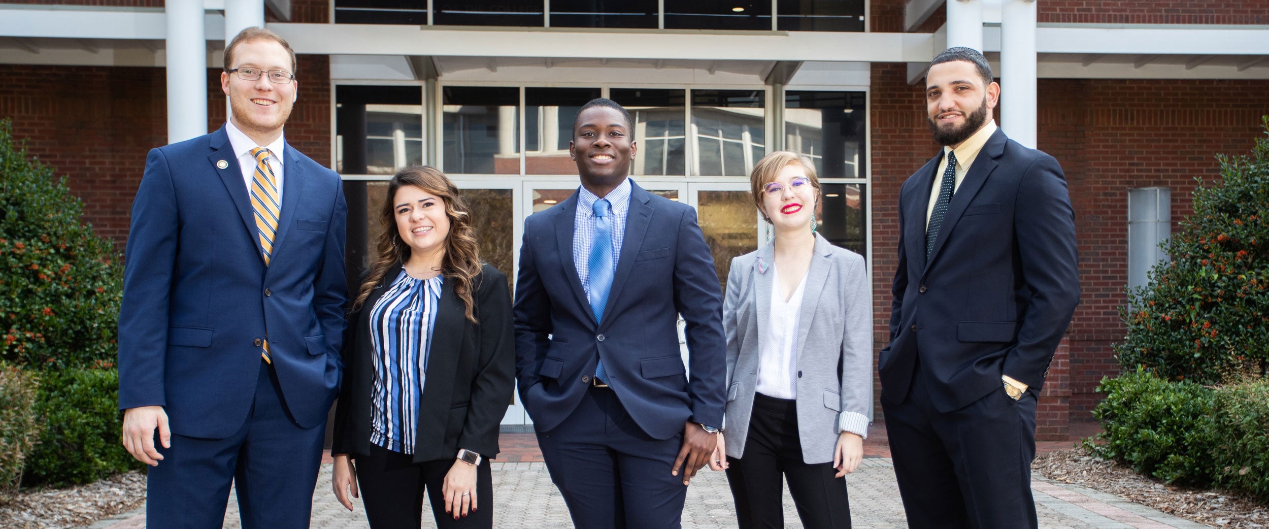 Five BBA in Economics students in business dress smile and stand in a row on one of Georgia Southern's campuses