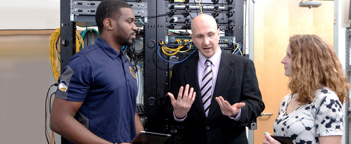 Standing in front of computer networking equipment, a professor explains a concept to two students enrolled in Georgia Southern's B.S. in Information Technology program