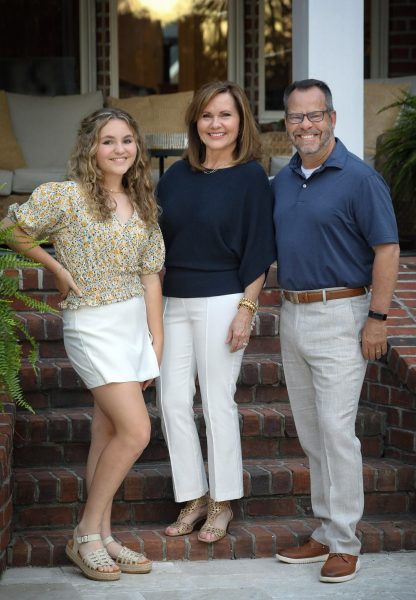 A family of three poses on brick steps leading up to a covered porch, presumably their home.