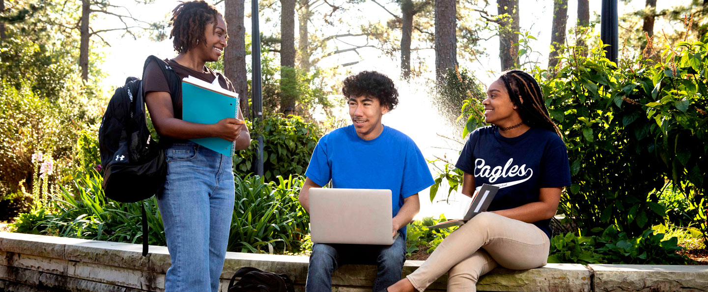 Three Georgia Southern students holding laptops or notebooks have a conversation outdoors near some greenery