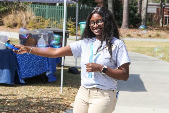 A young woman wearing glasses and a gray T-shirt with a printed design smiles while playing with a bubble wand outdoors.