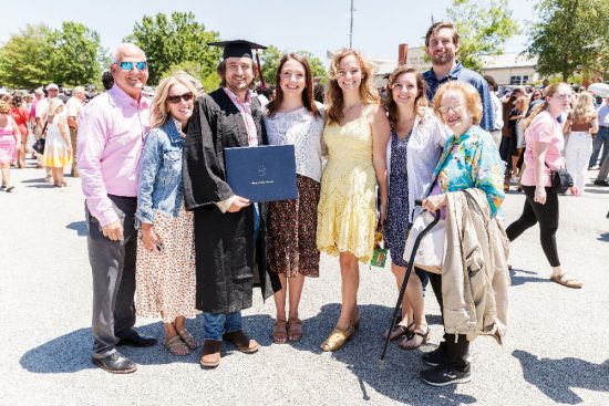 A group of eight people, including a Georgia Southern graduate in a black cap and gown, pose together outdoors on a sunny day. The graduate holds a diploma, surrounded by smiling family and friends, dressed in casual and summery outfits.