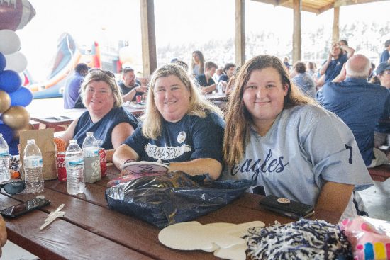 Three women sit together at a picnic table under a pavilion, wearing Georgia Southern Eagles gear. They smile at the camera with snacks and drinks on the table in front of them. In the background, other event attendees are visible, along with an inflatable slide.