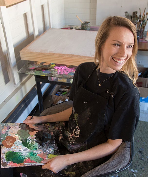 A Georgia Southern art student wearing an apron smiles while looking away from a palette holding multiple paint colors