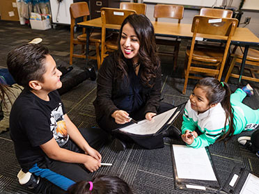 A student teacher from Georgia Southern's Department of Curriculum, Foundations and Reading and a group of students sitting on a rug discuss a worksheet.