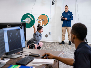 An athlete lifts a barbell while two students from Georgia Southern's M.S. in Kinesiology, Sport and Exercise Psychology program record their performance with a camera and computer