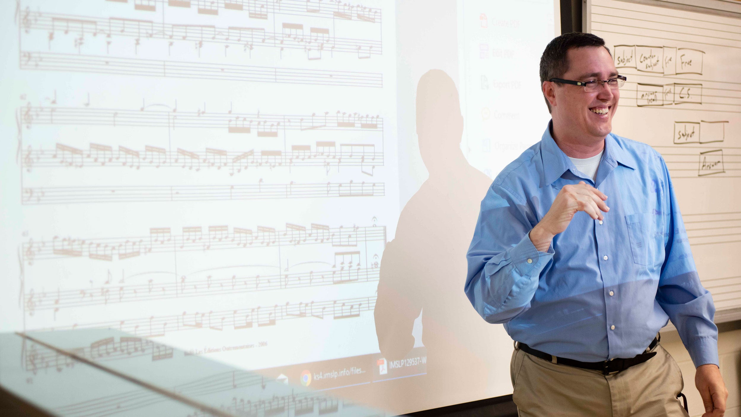 Georgia Southern professor smiling in front of a classroom with a white board behind him that has musical notes projected on it.