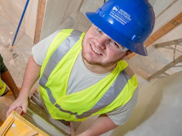 A Georgia Southern B.S. in Civil Engineering student wearing a hard hat and hi-viz vest looks upward while standing on a ladder