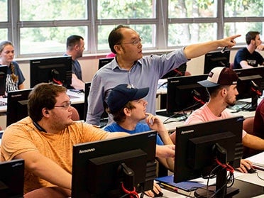 Sitting behind computers, Georgia Southern B.S. in Electrical Engineering students turn their heads to look where a faculty member is pointing