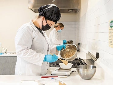 In a lab, a Georgia Southern B.S. in Nutrition and Food Science student spoons a mixture from a large metal bowl into a smaller bowl placed on a scale