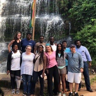 Study abroad group poses in front of a stunning waterfall. The flag of Ghana can be seen in the background.