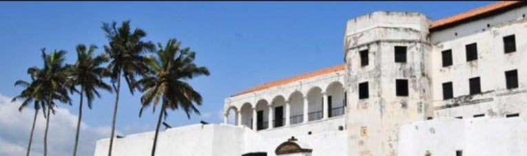 A group of palm trees stands starkly in front of a whitewashed fortress with terracotta tiled roof, a turret section and a columned balcony.